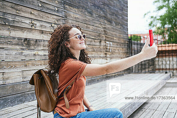 Smiling young woman wearing backpack taking selfie through smart phone