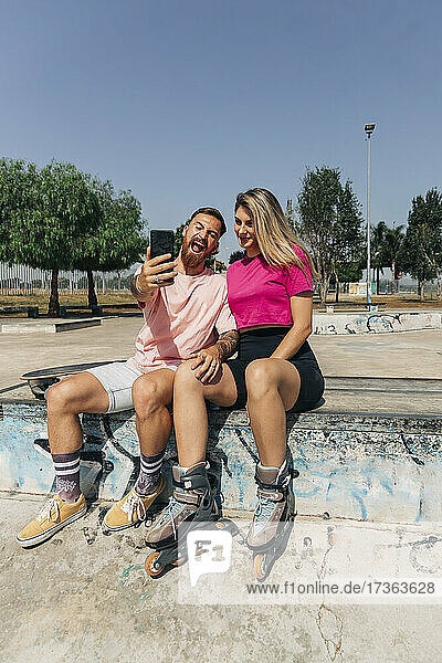 Playful young man taking selfie with girlfriend while sitting at skateboard park on sunny day