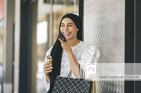 Happy woman with ice cream talking on smart phone in front of window