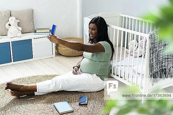 Lächelnde schwangere junge Frau  die ein Selfie macht  während sie vor der Krippe zu Hause sitzt