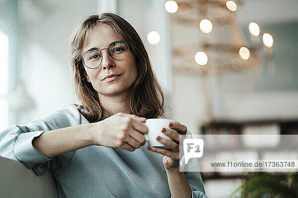 Junge Frau mit Brille hält eine Kaffeetasse  während sie in einem Café sitzt