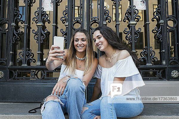Lächelnde Freundinnen machen ein Selfie  während sie vor einem Metalltor sitzen