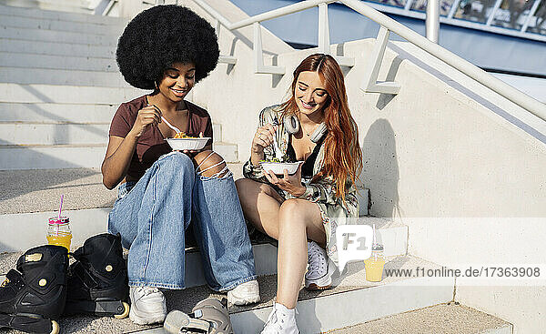 Smiling Afro woman eating food with female friend on steps during sunny day