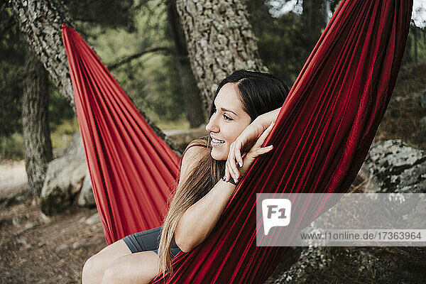 Lächelnde junge Frau  die auf einer Hängematte im Wald sitzt und nachdenkt