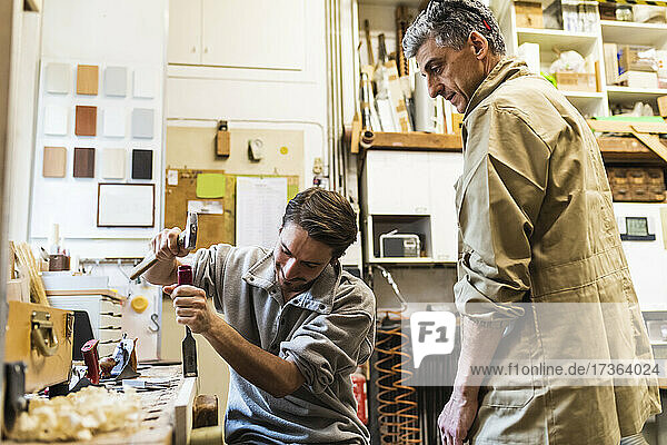 Male craftsperson hammering wood while owner looking in workshop