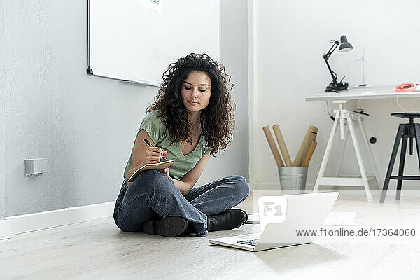 Geschäftsfrau schreibt auf Papier  während sie im Schneidersitz auf dem Boden im Büro sitzt