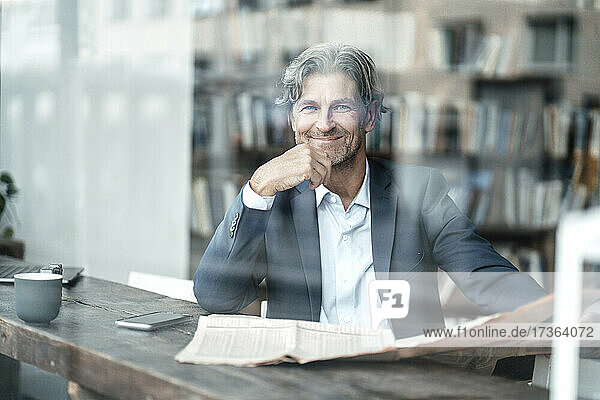 Lächelnder Geschäftsmann mit Zeitung im Café sitzend durch Glasfenster gesehen