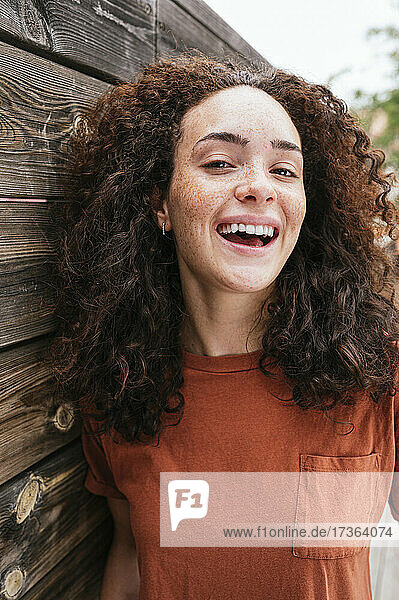 Fröhliche junge Frau mit lockigem Haar an einer Holzwand