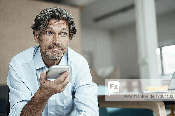 Älterer Geschäftsmann  der im Büro sitzend mit seinem Smartphone telefoniert