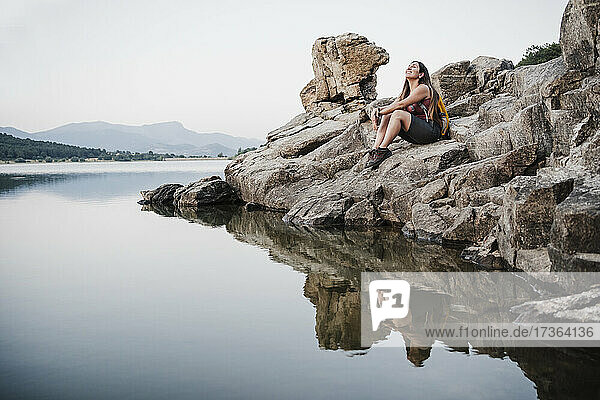 Lächelnde junge Frau mit Rucksack und Blick nach oben  während sie auf einem Felsen in der Nähe eines Sees sitzt