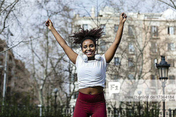 Fröhliche junge Sportlerin mit erhobener Hand beim Springen im Park
