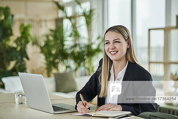 Glückliche blonde Teenager-Praktikantin  die wegschaut  während sie mit einem Tagebuch am Schreibtisch im Büro sitzt