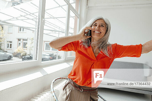 Lächelnde Geschäftsfrau  die mit einem Smartphone spricht  während sie im Büro mit einem Hula-Hoop-Reifen übt