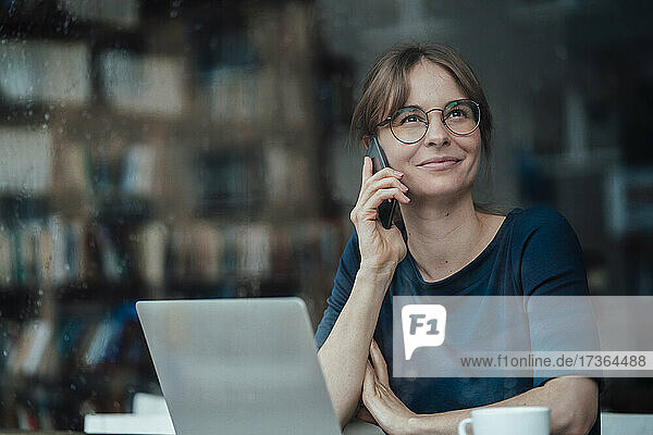 Lächelnde weibliche Fachkraft  die wegschaut und mit einem Smartphone in einem Café spricht  gesehen durch ein Glasfenster