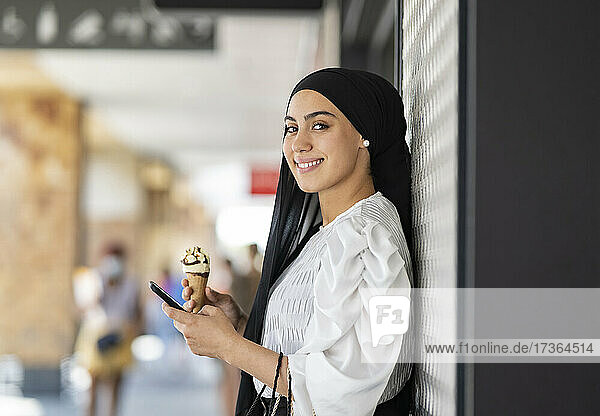 Smiling woman in hijab holding mobile phone while having ice cream