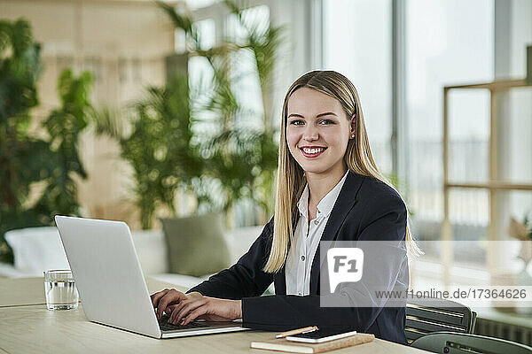 Smiling blond female trainee sitting with laptop at desk in office
