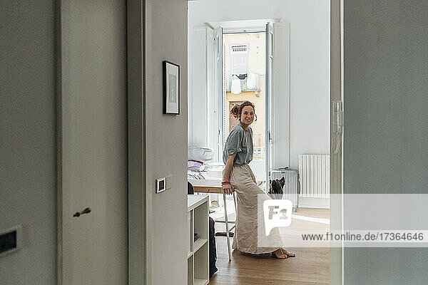 Smiling woman sitting on table looking through doorway in apartment
