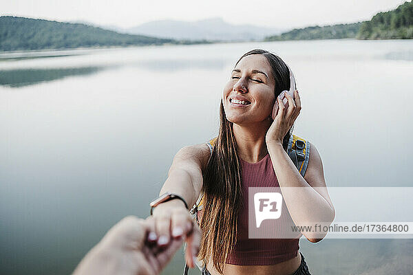 Lächelnde junge Frau mit geschlossenen Augen hält die Hand eines Freundes am Seeufer