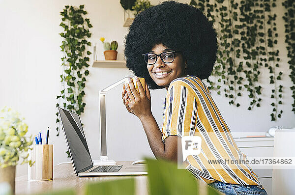 Lächelnde Afro-Frau  die eine Kaffeetasse hält  während sie am Schreibtisch sitzt