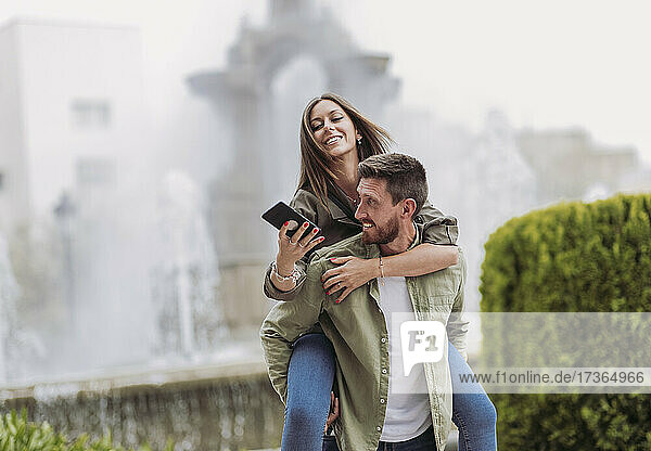 Lächelnde Frau  die ein Mobiltelefon benutzt  während sie auf ihrem Freund huckepack sitzt