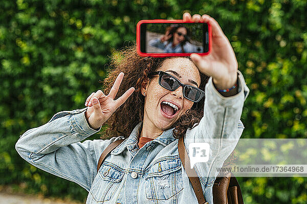 Junge Frau  die ein Selfie mit ihrem Smartphone macht  während sie das Friedenszeichen mit offenem Mund zeigt