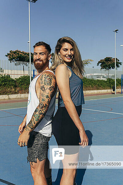 Glückliches junges Paar steht Rücken an Rücken am Basketballplatz an einem sonnigen Tag