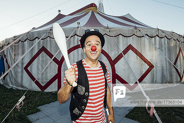 Lächelnder männlicher Clown  der einen Jonglierstift hält  während er vor einem Zirkuszelt steht
