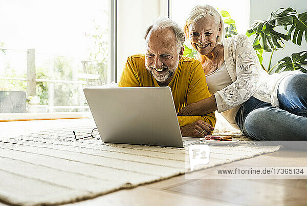 Lächelndes Paar,  das einen Laptop benutzt,  während es sich auf einem Teppich zu Hause entspannt