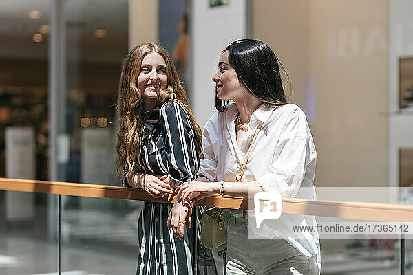 Smiling female friends looking at each other while leaning on railing at shopping mall