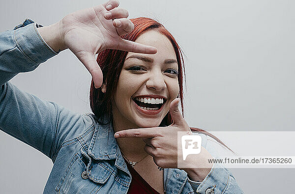 Lächelnde junge Frau macht Fingerrahmen vor weißem Hintergrund