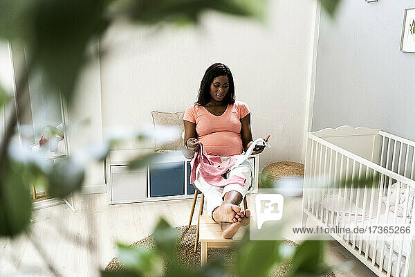 Schwangere Frau sitzt mit Babykleidung auf einem Stuhl im Schlafzimmer
