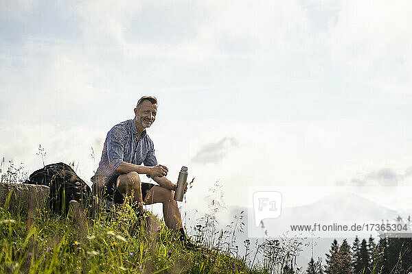 Lächelnder Mann mit isoliertem Getränkebehälter auf einem Berg sitzend