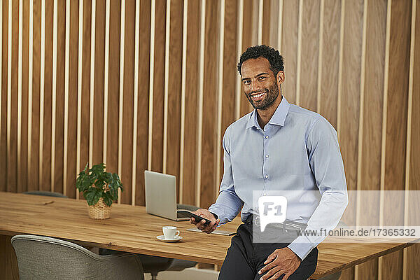 Männlicher Berufstätiger  der ein Mobiltelefon hält und sich auf einen Konferenztisch stützt