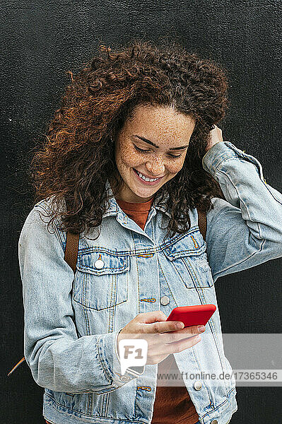 Smiling young woman text messaging through smart phone while standing with hand in hair