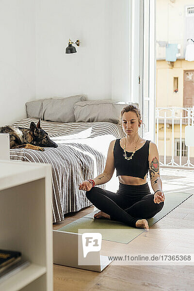 Frau  die im Schlafzimmer sitzend mit dem Laptop Yoga lernt