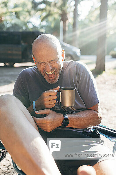 Unbekümmerter Mann mit Kaffeetasse lachend an einem sonnigen Tag im Urlaub