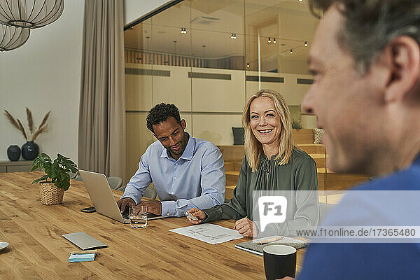 Lächelnde Geschäftsfrau  die einen Kollegen ansieht  während sie neben einem Geschäftsmann im Sitzungssaal sitzt