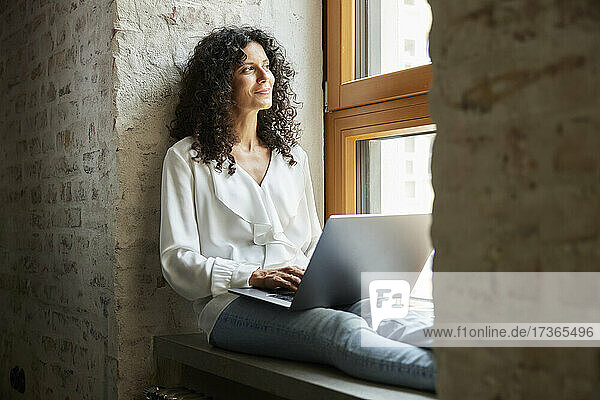 Geschäftsfrau  die mit ihrem Laptop auf der Fensterbank sitzt und träumt