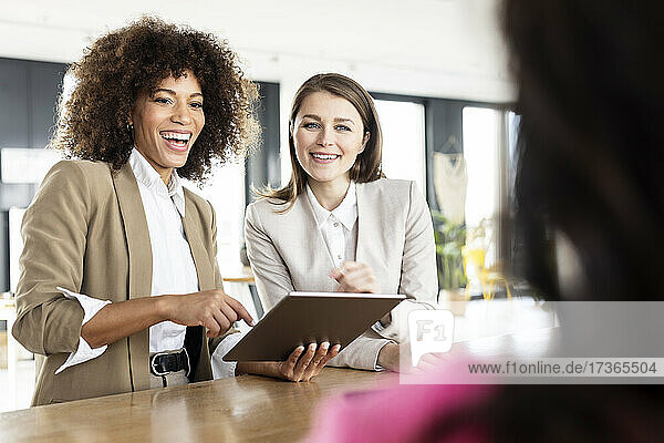 Glückliche weibliche Fachkräfte mit digitalem Tablet  die einen Kollegen bei einer Besprechung im Büro ansehen