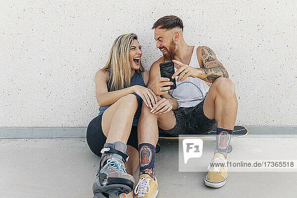 Fröhlicher Mann  der sein Smartphone hält und lachend mit seiner Freundin an der Wand lehnt