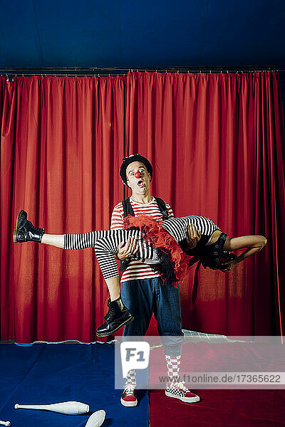 Männlicher Clown mit offenem Mund  der eine weibliche Darstellerin auf der Bühne eines Zirkus' trägt