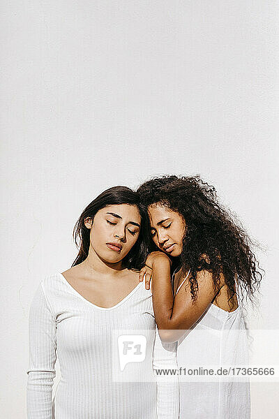 Female friends with eyes closed leaning on each other in front of white wall