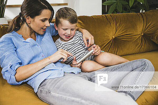 Frau sitzt mit ihrem Sohn auf dem heimischen Sofa und benutzt ein Smartphone