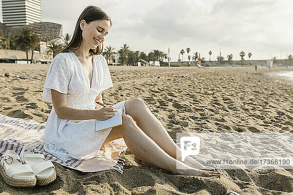Lächelnde Frau  die auf einem Handtuch sitzend in ein Buch schreibt