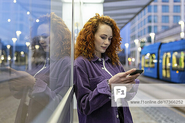 Junge Frau benutzt ein Mobiltelefon  während sie sich an einer Straßenbahnhaltestelle an ein Glas lehnt