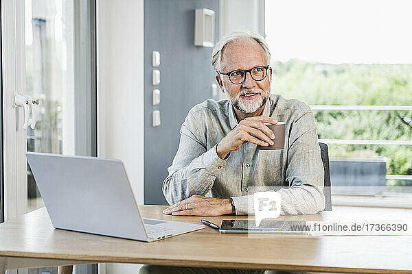 Businessman looking away holding while coffee mug at desk in home office