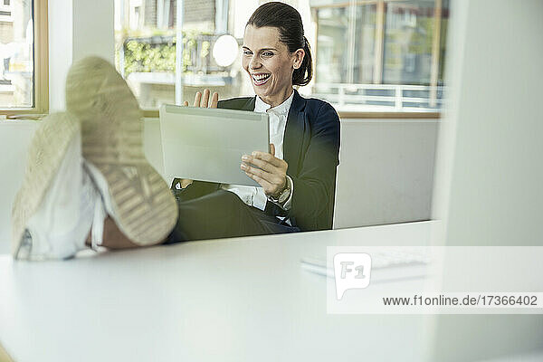 Fröhliche Geschäftsfrau winkt mit der Hand zu einem Videoanruf über ein digitales Tablet im Büro