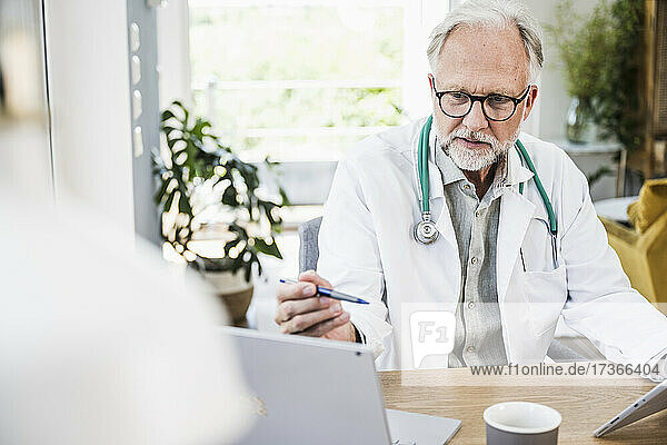 Reifer männlicher Arzt  der auf einen Laptop schaut und einen Stift am Schreibtisch hält