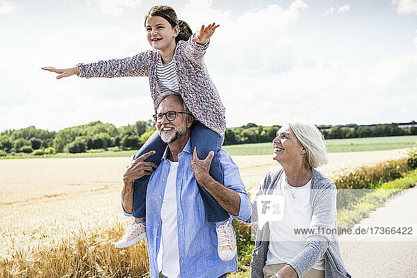 Smiling girl with arms outstretched sitting on man shoulder during sunny day