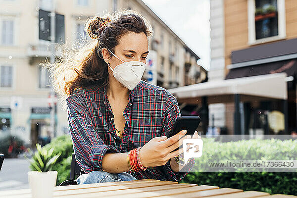 Woman with protective face mask using mobile phone at sidewalk cafe on sunny day